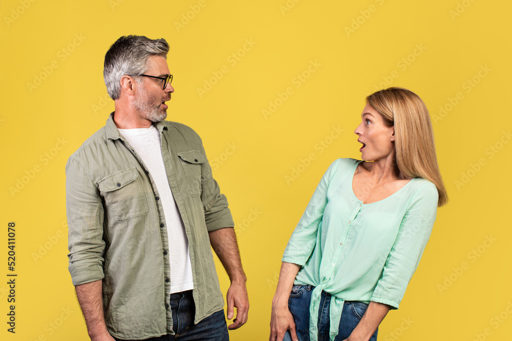 Fototapeta premium Shocked middle aged woman and her husband looking at each other with open mouths, feeling surprised on yellow background