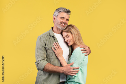 Portrait of loving middle aged spouses hugging with closed eyes while standing isolated over yellow studio background