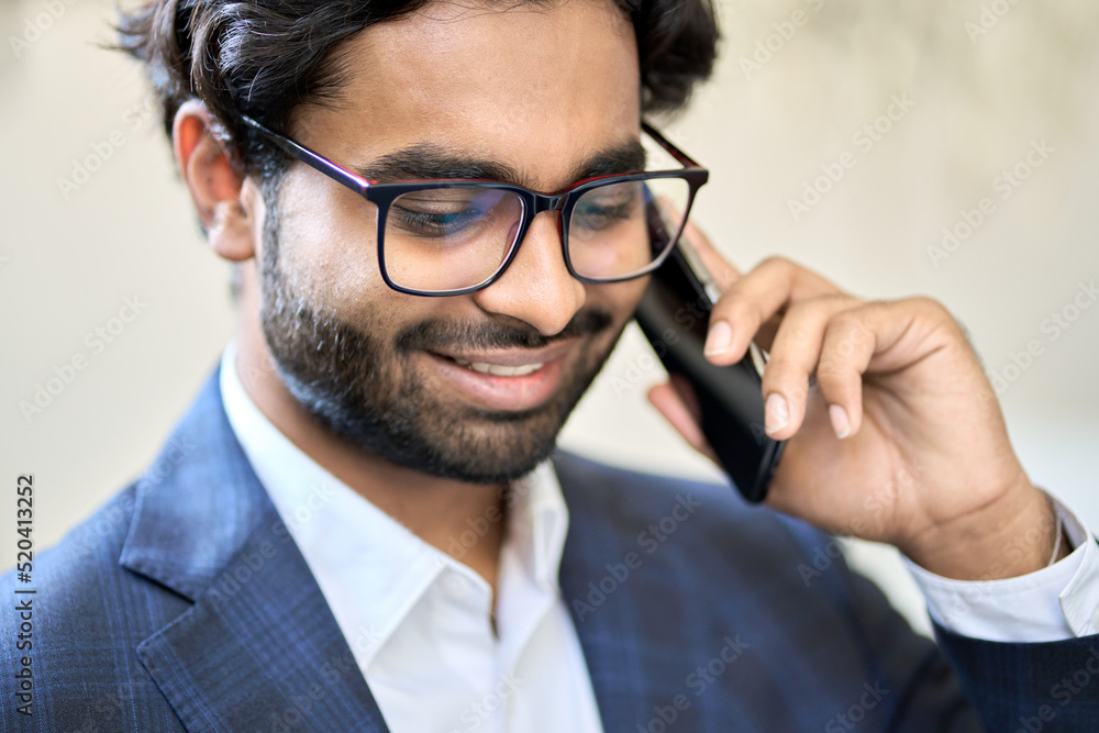 Smiling young indian business man ceo manager wearing suit and glasses holding smartphone talking on cell phone making call in office working consulting client providing business service by cellphone.
