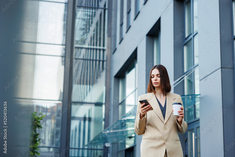 A young brunette girl against the backdrop of a business center, office center. On the go, he reads the news from his smartphone.