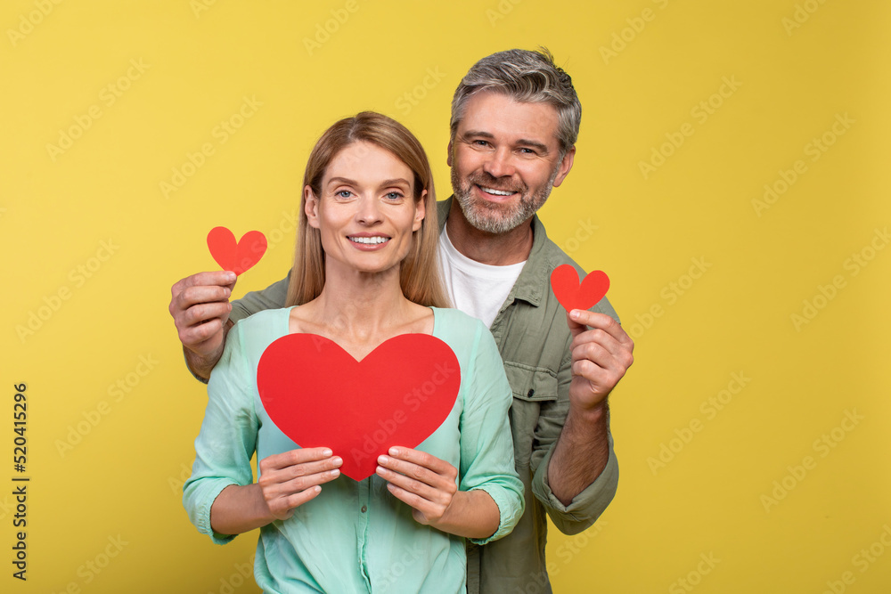 Love is in the air. Loving middle aged spouses with red paper hearts in hands posing over yellow studio background