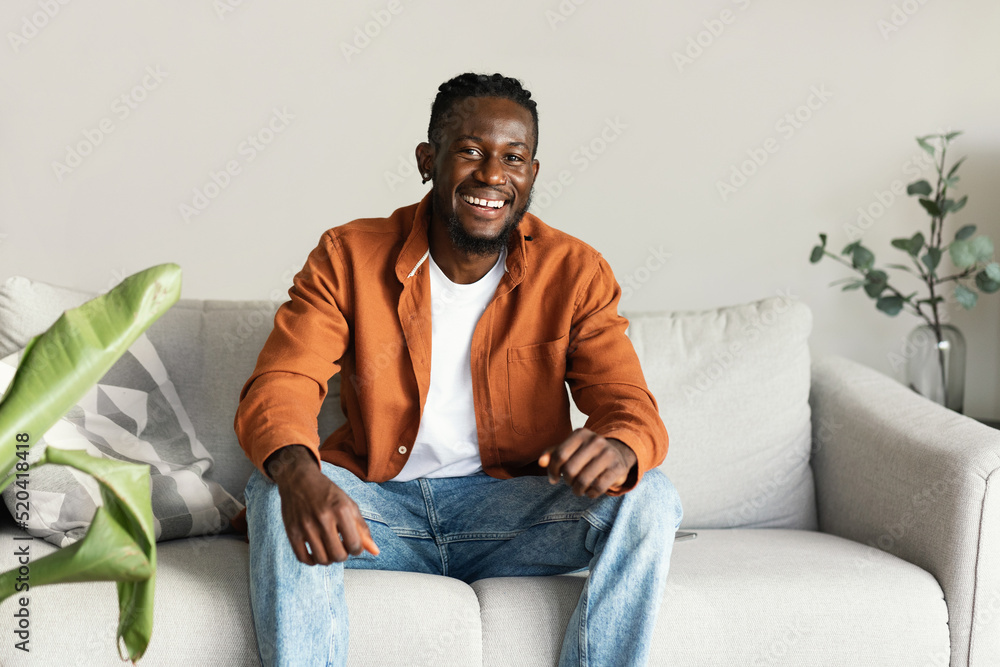 Portrait of handsome african american man smiling and looking at camera, sitting on sofa at home, free space