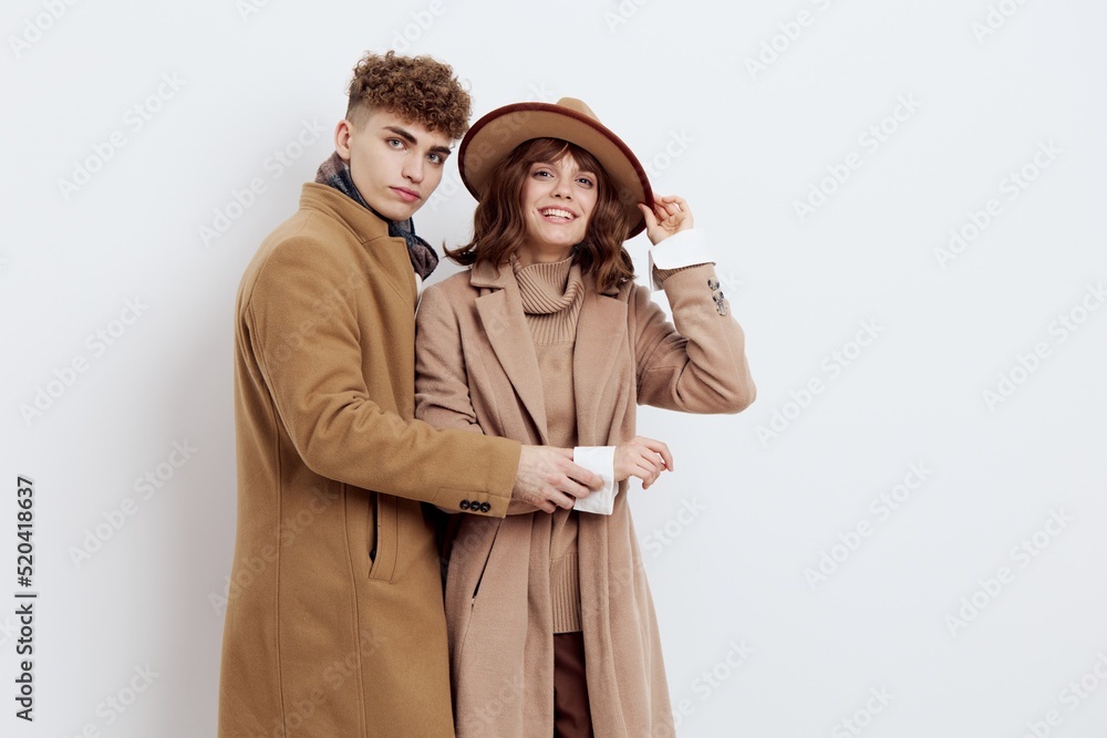 a cute couple stands on a light background dressed in coats and sweaters, a man looks tenderly at a woman and hugs her to him while she adjusts her hat with one hand a cute couple stands on a light background dressed in coats and sweaters, a man looks tenderly at a woman and hugs her to him while she adjusts her hat with one hand