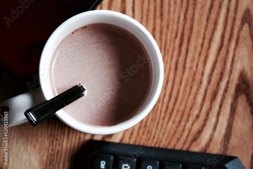 Hot chocolate in a cup placed on a wooden table with a computer and a mobile phone.