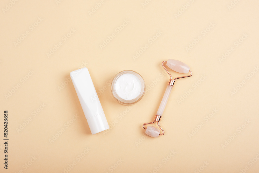 Rose quartz crystal facial roller, cream jar and tube on neutral beige background. Natural cosmetics, skin care concept. Top view, flat lay, mockup Rose quartz crystal facial roller, cream jar and tube on neutral beige background. Natural cosmetics, skin care concept. Top view, flat lay, mockup
