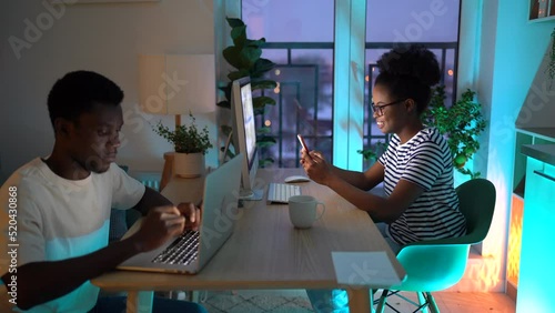 Cheerful woman shows photos on smartphone to man trying to distract from writing article on laptop. Young African American couple works on computers sitting at wooden table in kitchen in late evening
