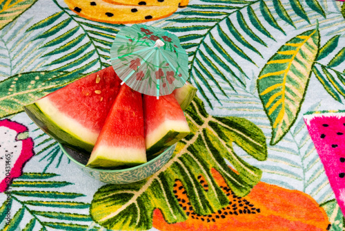 Sliced watermelon with a small red umbrella  on a tropical background of palm leaves.
