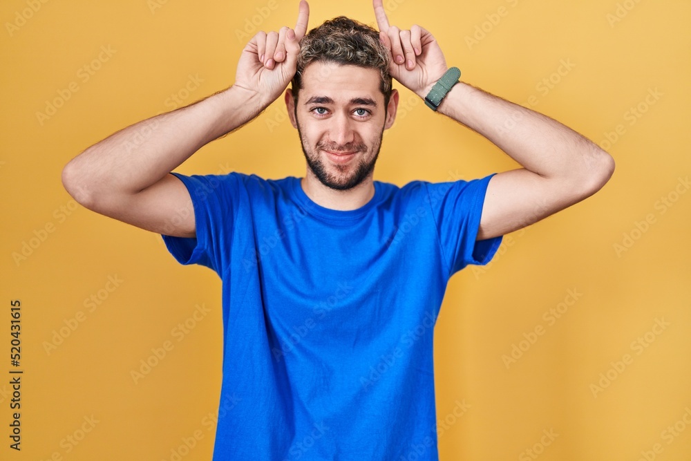 Hispanic man with beard standing over yellow background doing funny gesture with finger over head as bull horns Hispanic man with beard standing over yellow background doing funny gesture with finger over head as bull horns