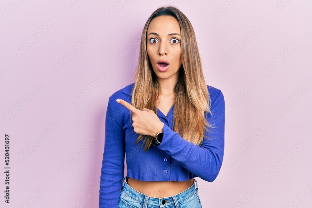 Beautiful hispanic woman wearing casual blue shirt surprised pointing with finger to the side, open mouth amazed expression. Beautiful hispanic woman wearing casual blue shirt surprised pointing with finger to the side, open mouth amazed expression.