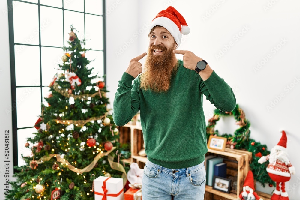 Redhead man with long beard wearing christmas hat by christmas tree smiling cheerful showing and pointing with fingers teeth and mouth. dental health concept. Redhead man with long beard wearing christmas hat by christmas tree smiling cheerful showing and pointing with fingers teeth and mouth. dental health concept.