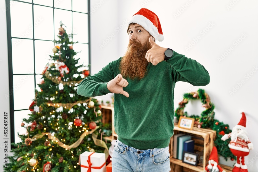 Redhead man with long beard wearing christmas hat by christmas tree doing thumbs up and down, disagreement and agreement expression. crazy conflict Redhead man with long beard wearing christmas hat by christmas tree doing thumbs up and down, disagreement and agreement expression. crazy conflict