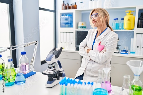 Middle age blonde woman working at scientist laboratory looking to the side with arms crossed convinced and confident