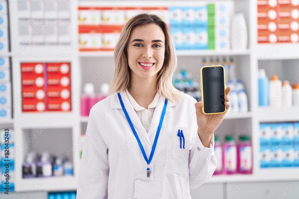 Young beautiful woman working at pharmacy drugstore showing smartphone screen looking positive and happy standing and smiling with a confident smile showing teeth