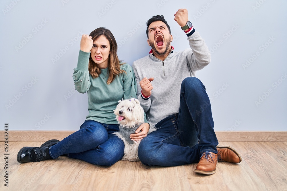 Young hispanic couple sitting on the floor with dog angry and mad raising fist frustrated and furious while shouting with anger. rage and aggressive concept. Young hispanic couple sitting on the floor with dog angry and mad raising fist frustrated and furious while shouting with anger. rage and aggressive concept.