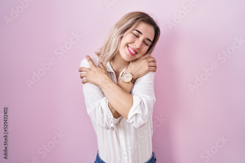 Young beautiful woman standing over pink background hugging oneself happy and positive, smiling confident. self love and self care