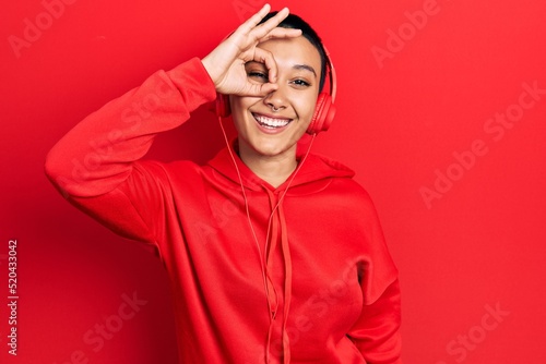 Beautiful hispanic woman with short hair listening to music using headphones doing ok gesture with hand smiling, eye looking through fingers with happy face. © Krakenimages.com