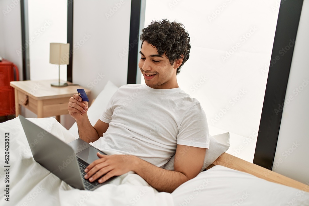 Fototapeta premium Young hispanic man buying using laptop and credit card sitting on the bed at bedroom.