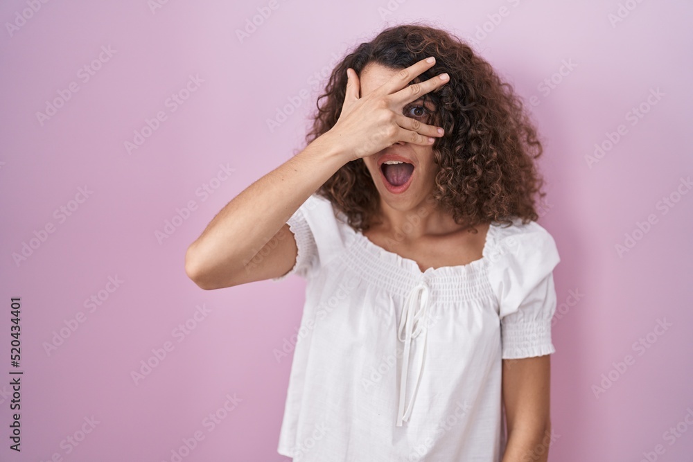 Hispanic woman with curly hair standing over pink background peeking in shock covering face and eyes with hand, looking through fingers with embarrassed expression. Hispanic woman with curly hair standing over pink background peeking in shock covering face and eyes with hand, looking through fingers with embarrassed expression.