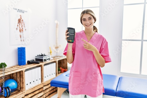 Young physiotherapist woman working at pain recovery clinic holding dataphone smiling happy pointing with hand and finger