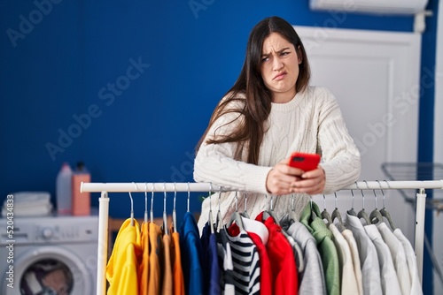 Young brunette woman waiting for laundry using smartphone clueless and confused expression. doubt concept.