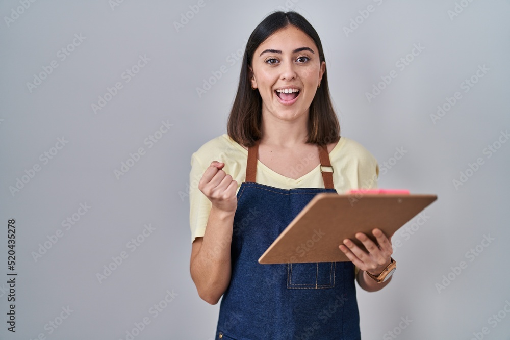 Hispanic girl wearing apron holding clipboard screaming proud, celebrating victory and success very excited with raised arms Hispanic girl wearing apron holding clipboard screaming proud, celebrating victory and success very excited with raised arms