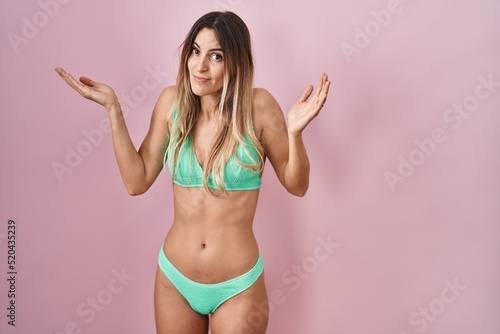 Young hispanic woman wearing bikini over pink background clueless and confused expression with arms and hands raised. doubt concept.