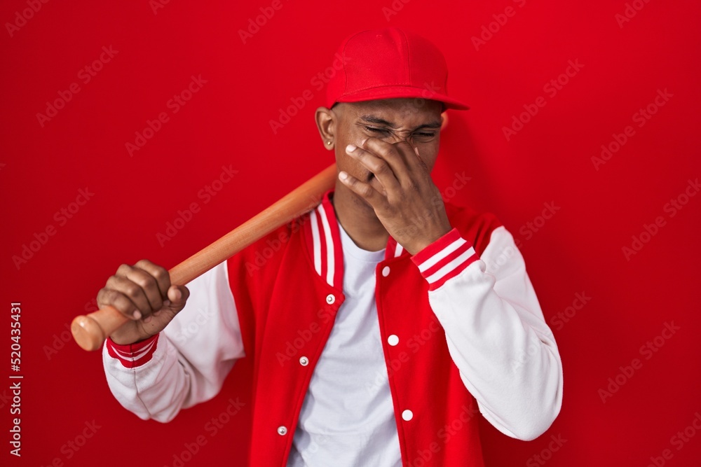 Young hispanic man playing baseball holding bat smelling something stinky and disgusting, intolerable smell, holding breath with fingers on nose. bad smell Young hispanic man playing baseball holding bat smelling something stinky and disgusting, intolerable smell, holding breath with fingers on nose. bad smell