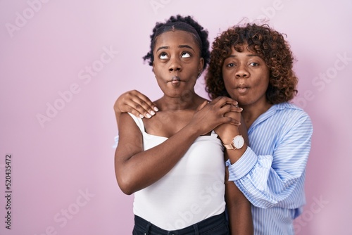 Two african women standing over pink background making fish face with mouth and squinting eyes, crazy and comical.