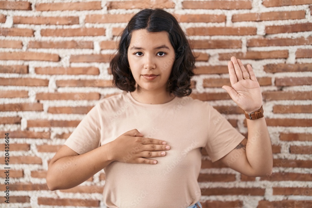 Young hispanic woman standing over bricks wall swearing with hand on chest and open palm, making a loyalty promise oath Young hispanic woman standing over bricks wall swearing with hand on chest and open palm, making a loyalty promise oath