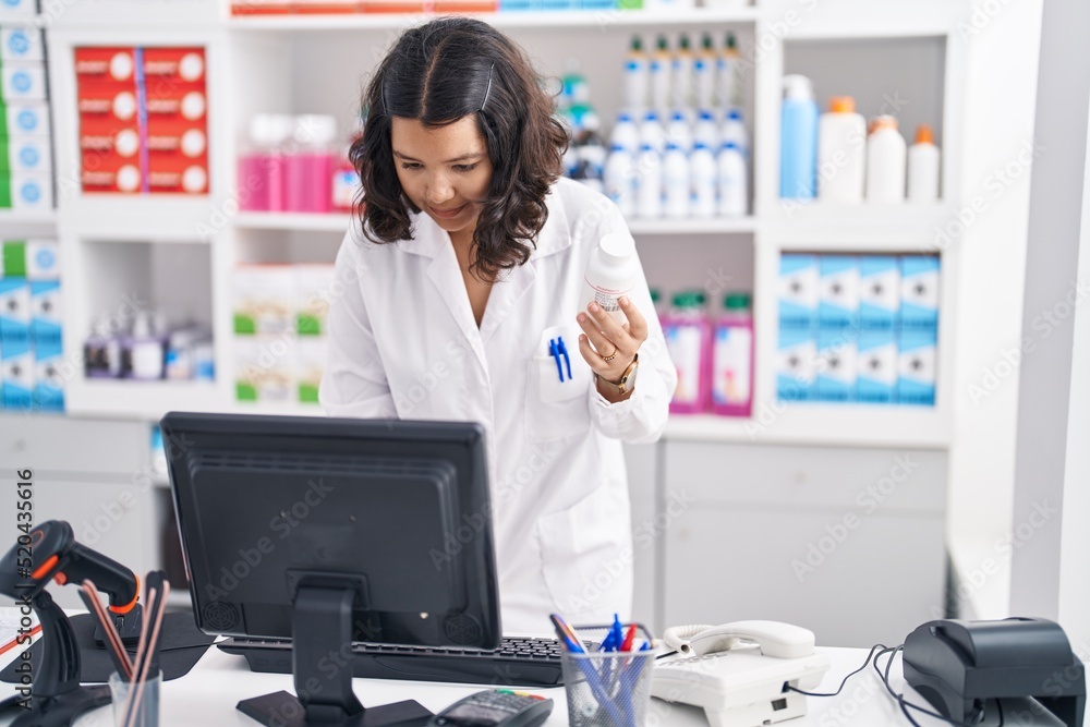 Young woman pharmacist holding pills bottle using computer at pharmacy Young woman pharmacist holding pills bottle using computer at pharmacy