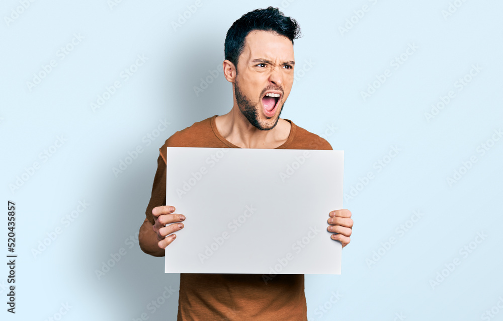 Hispanic man with beard holding blank empty banner angry and mad screaming frustrated and furious, shouting with anger. rage and aggressive concept. Hispanic man with beard holding blank empty banner angry and mad screaming frustrated and furious, shouting with anger. rage and aggressive concept.