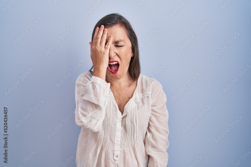 Middle age hispanic woman standing over blue background yawning tired covering half face, eye and mouth with hand. face hurts in pain. Middle age hispanic woman standing over blue background yawning tired covering half face, eye and mouth with hand. face hurts in pain.