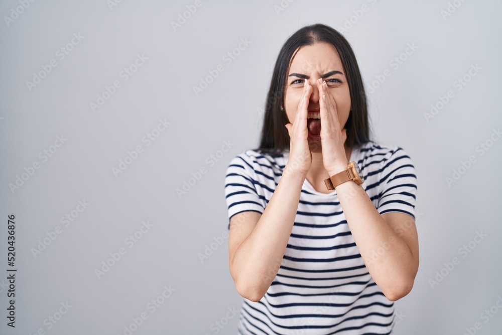 Young brunette woman wearing striped t shirt shouting angry out loud with hands over mouth Young brunette woman wearing striped t shirt shouting angry out loud with hands over mouth
