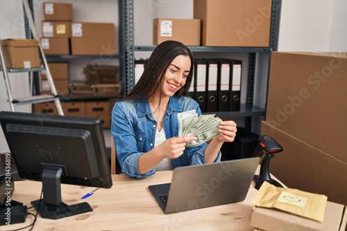 Young beautiful hispanic woman ecommerce business worker using laptop holding dollars at office