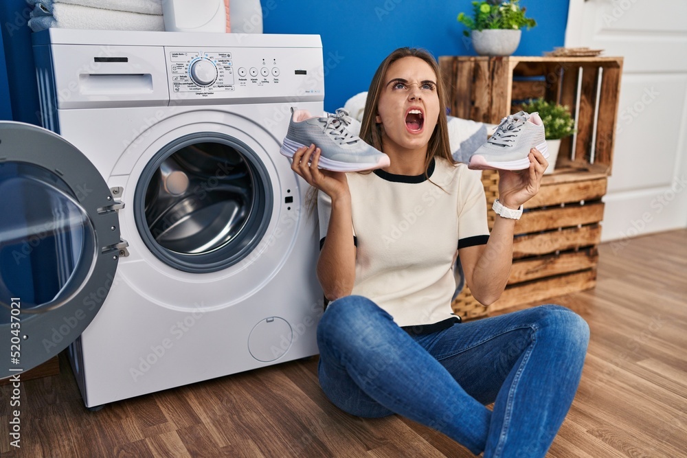 Young woman putting sneakers in washing machine angry and mad screaming frustrated and furious, shouting with anger looking up. Young woman putting sneakers in washing machine angry and mad screaming frustrated and furious, shouting with anger looking up.