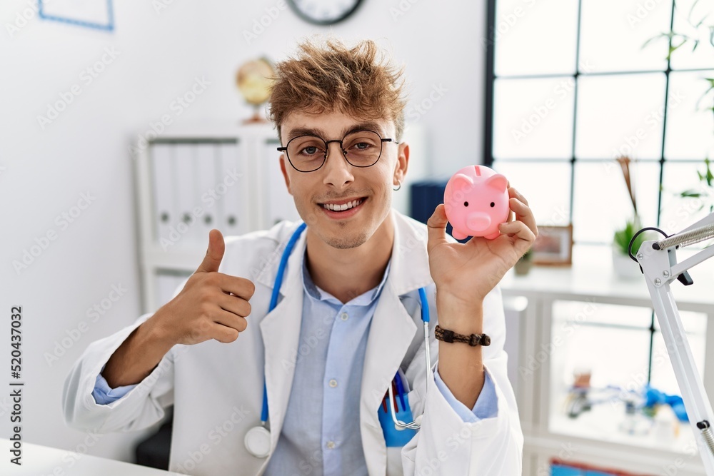 Young caucasian doctor man wearing doctor uniform holding piggy bank at the clinic smiling happy and positive, thumb up doing excellent and approval sign Young caucasian doctor man wearing doctor uniform holding piggy bank at the clinic smiling happy and positive, thumb up doing excellent and approval sign