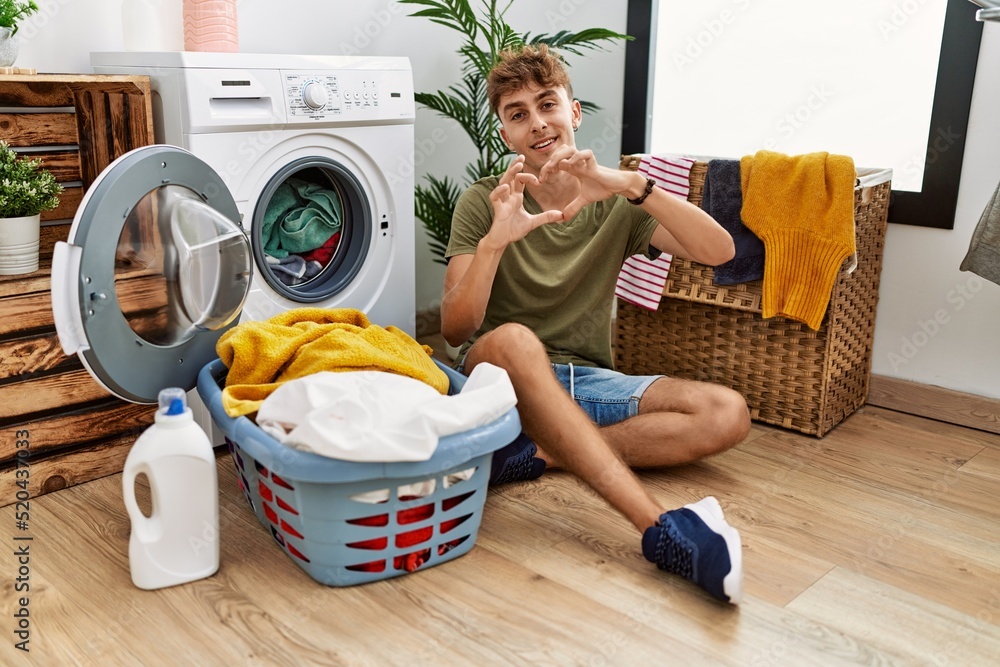 Young caucasian man putting dirty laundry into washing machine smiling in love doing heart symbol shape with hands. romantic concept. Young caucasian man putting dirty laundry into washing machine smiling in love doing heart symbol shape with hands. romantic concept.