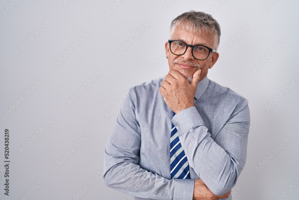 Hispanic business man with grey hair wearing glasses looking confident at the camera smiling with crossed arms and hand raised on chin. thinking positive. Hispanic business man with grey hair wearing glasses looking confident at the camera smiling with crossed arms and hand raised on chin. thinking positive.
