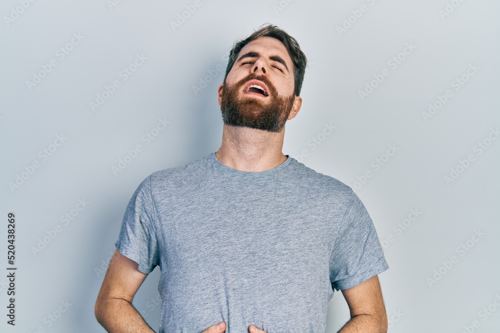 Caucasian man with beard wearing casual grey t shirt with hand on stomach because nausea, painful disease feeling unwell. ache concept. Caucasian man with beard wearing casual grey t shirt with hand on stomach because nausea, painful disease feeling unwell. ache concept.