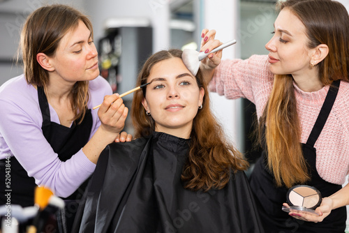Two professional female makeup artists working in a beauty salon apply makeup on the face of a young woman