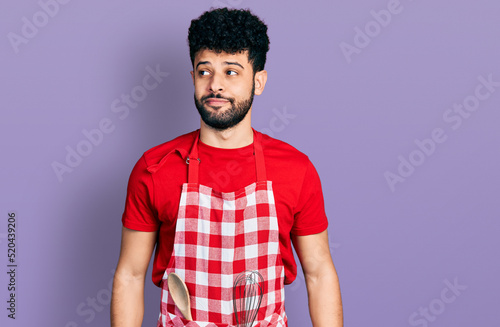 Young arab man with beard wearing cook apron smiling looking to the side and staring away thinking.