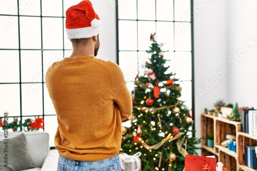 Young arab man on back view standing with arms crossed gesture by christmas tree at home. photo