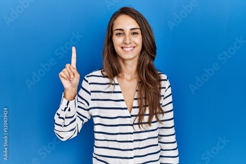 Young hispanic woman standing over blue isolated background showing and pointing up with finger number one while smiling confident and happy.