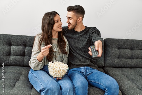 Young hispanic couple watching film and eating popcorn sitting on the sofa at home.