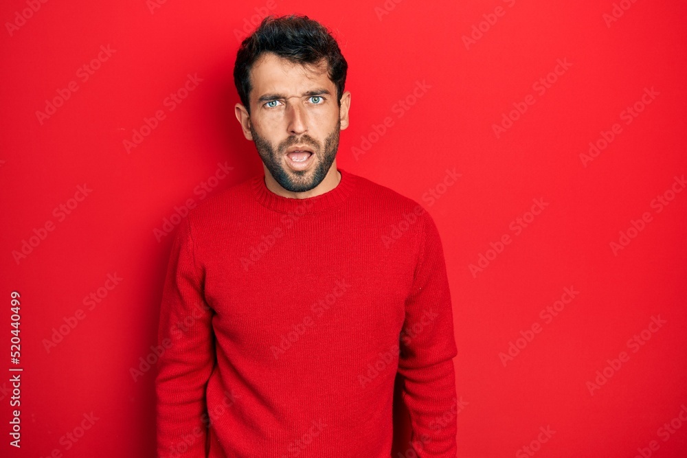 Handsome man with beard wearing casual red sweater in shock face, looking skeptical and sarcastic, surprised with open mouth Handsome man with beard wearing casual red sweater in shock face, looking skeptical and sarcastic, surprised with open mouth