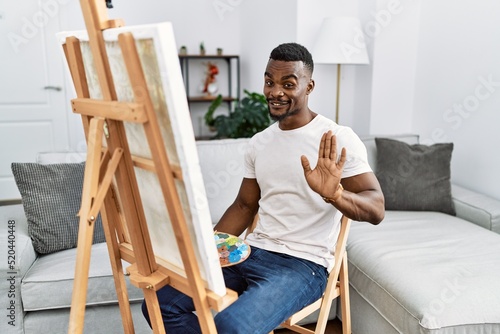 Young african man painting on canvas at home waiving saying hello happy and smiling, friendly welcome gesture