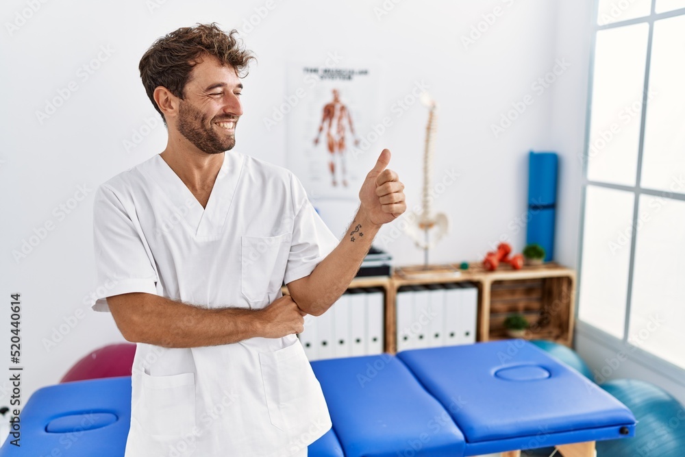 Young handsome physiotherapist man working at pain recovery clinic looking proud, smiling doing thumbs up gesture to the side