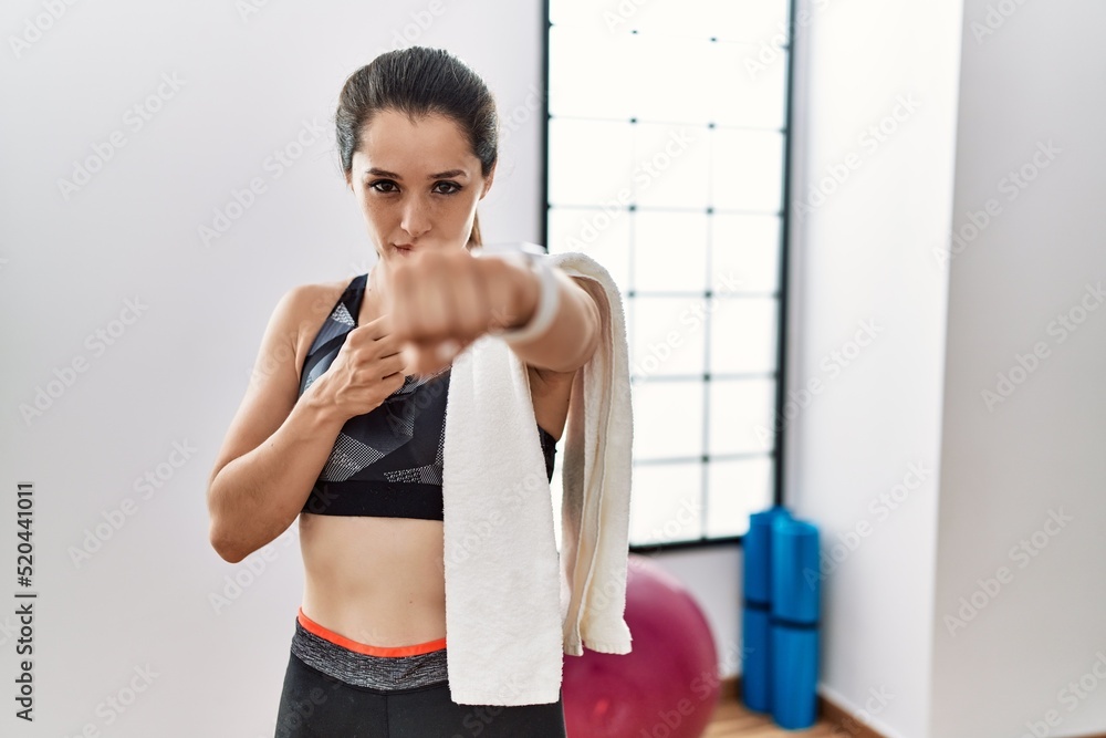 Young brunette woman wearing sportswear and towel at the gym punching fist to fight, aggressive and angry attack, threat and violence Young brunette woman wearing sportswear and towel at the gym punching fist to fight, aggressive and angry attack, threat and violence