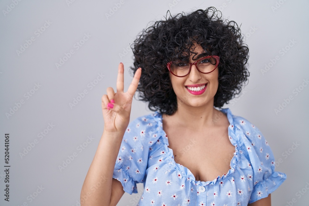 Young brunette woman with curly hair wearing glasses over isolated background showing and pointing up with fingers number two while smiling confident and happy. Young brunette woman with curly hair wearing glasses over isolated background showing and pointing up with fingers number two while smiling confident and happy.