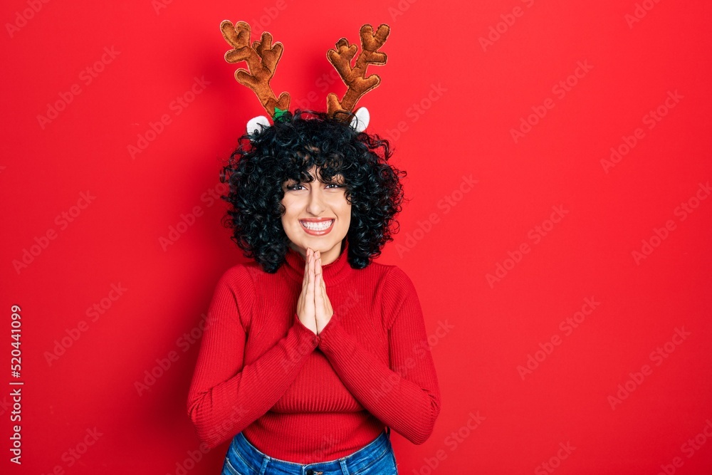 Young middle east woman wearing cute christmas reindeer horns praying with hands together asking for forgiveness smiling confident. Young middle east woman wearing cute christmas reindeer horns praying with hands together asking for forgiveness smiling confident.